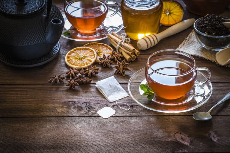 Filled tea cup surrounded by a teaspoon, teapot, teabag and some ingredientes like dried black tea leaves, anise, dried orange, cinnamon sticks and a honey jar.
